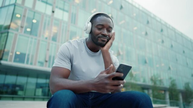 African american man music lover in headphones enjoying listening music on smartphone sitting on city street outdoors. Positive smiling guy melomaniac rhythmically nodding head relaxing moving body.