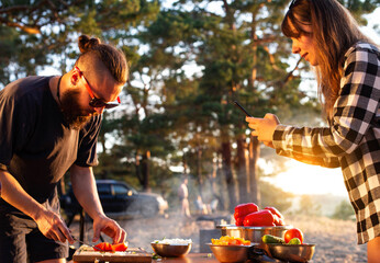 A brutal caucasian man with a beard and black glasses is preparing a vegetable salad in nature against the backdrop of an evening sunset. The girl shoots on the phone. Cooking in nature, tourism. 