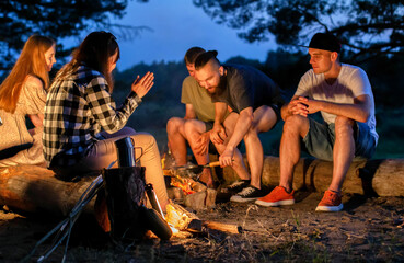 A company of young people sit around a fire at night on the banks of the river and relax in nature. Cooking food on a fire, outdoors - Powered by Adobe