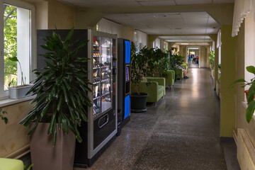 Side view of free standing coffee and combo food (snack, cold drink and food) vending machines standing in corridor with green sofas and plants in public clinic. Copy space for your text. - obrazy, fototapety, plakaty