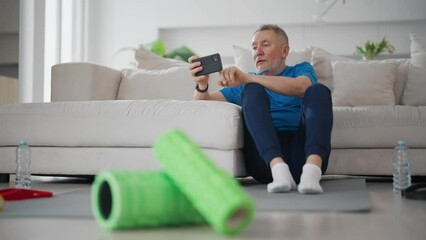 Senior man finds video training in smartphone for doing sport exercises at home sits on yoga mat in living room. Elderly male in sportswear prepares to workout. Sport active healthy lifestyle concept.