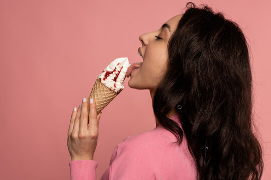 Side view of a woman licking a tasty vanilla ice cream in the waffle cone during the studio photo shoot on the pink background