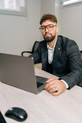 Vertical portrait of confident businessman wearing stylish suit and eyeglasses working on laptop computer sitting at office desk. Successful bearded CEO looking at camera with serious expression.