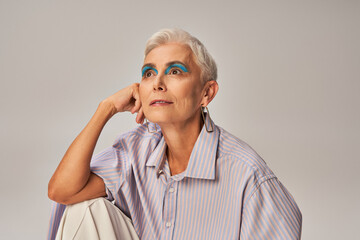 pensive and stylish senior woman in blue striped shirt and bold makeup looking away on grey