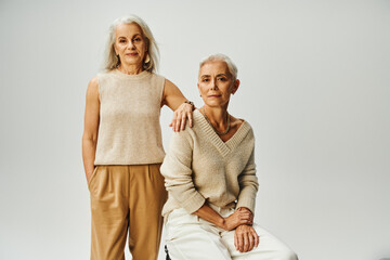 trendy mature woman standing with hand in pocket near elegant female friend sitting on grey backdrop