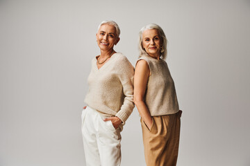 smiling senior women in fashionable clothes standing back to back with hands in pockets on grey