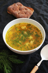 Chicken soup with dill in a white plate next to a spoon, bread and dill on a dark background