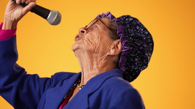 Closeup funny crazy toothless grandmother with a fashionable look with glasses, boa, singing enthusiastically into a microphone and dancing isolated on solid yellow background