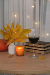 autumn home decoration. Vase autumn yellow leafes, glass of red wine, books, candle and pumpkin on round wooden table.