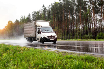 A small truck transports cargo against the backdrop of a forest on a wet road in the rain, industry