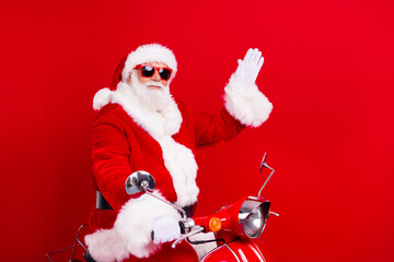 Portrait of positive optimistic santa claus with white beard in stylish sunglass waving palm sit on moped isolated on red color background