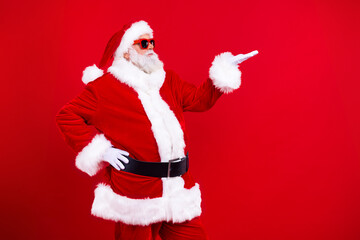 Portrait of positive santa claus with white beard in sunglass look at arm hold product empty space isolated on red color background