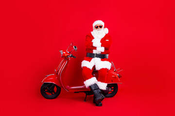 Portrait of satisfied santa claus with white beard sitting on red vintage scooter hold arms crossed isolated on red color background
