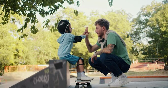 A little boy in a Blue sweater and a black safety helmet and His dad with curly hair in a Green T-shirt high-five each other on a skateboard in the park
