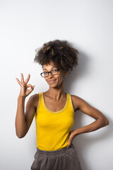 Young successful black casual woman wearing glasses and doing ok gesture. Afro hairstyle cheerful model against white wall.