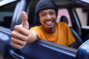Close-up of happy Asian online taxi driver showing thumbs up to camera while sitting in a car