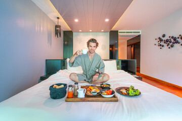 Handsome Man Savors a Big Room Service Breakfast in the Comfort of His Hotel Bed