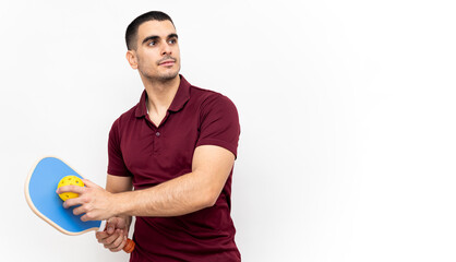 A handsome young man in sportswear poses on a white studio background with a pickleball racket and a ball. The man looks to the side. Pickle ball concept. Racquet sports. Copy space for advertising.
