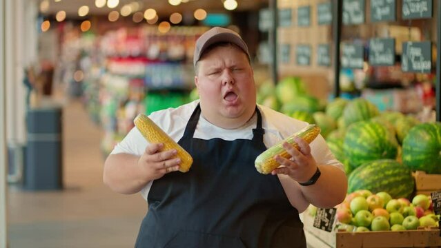 A cheerful overweight male supermarket worker in a white T-shirt and a black apron in a gray cap dances along with heads of corn and uses them as a microphone during his fun at work against the