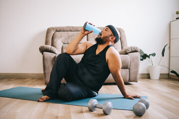 Overweight man is sitting on a yoga mat drinking water feeling exhausted after a hard training at...