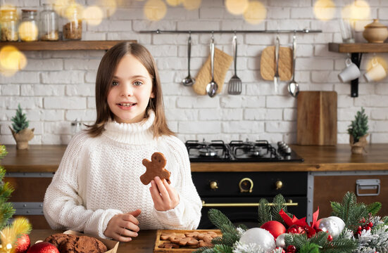 Child standing at wooden table in Christmas decorated kitchen. Kid holding gingerbread man, cookies on table. Bokeh framed image