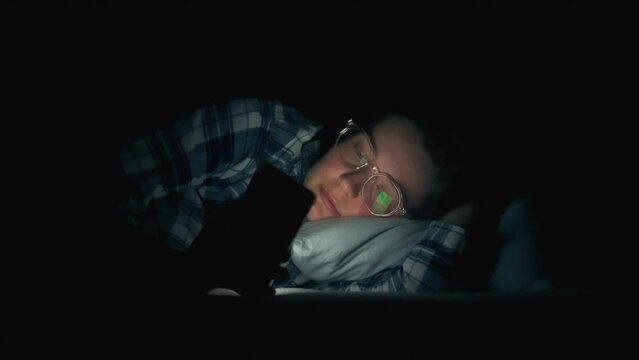 Mobile phone. Mature woman under a blanket does not sleep at night addicted to the phone in a dark room. News follower looking at the bright screen of a modern phone while studying trends, insomnia