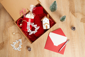 Gift wrapping for Christmas and New Year. A craft box with gifts: a red mug, knitted mittens, a wooden toy house and a Christmas tree toy. An empty piece of paper for congratulations in a red envelope