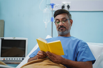 Wide shot Indian elderly senior man reading book at hospital bed after recovery - concept of...