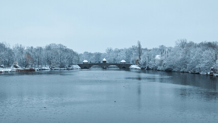 The Palmengarten weir in Leipzig in winter: Snow covers the bank, the frozen Elster, and light fog...