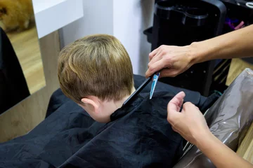 Fototapeten A little cute boy sits in a hairdresser's at the stylist's, a schoolchild is getting hair cut in a beauty salon, a child at a barbershop's, a short men's haircut © Miri García