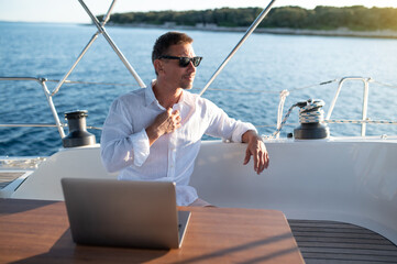 Man in white tshirt sitting on a yacht deck and typing on a laptop