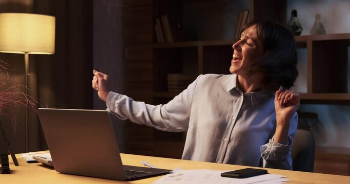 Contented woman lets loose and dances joyfully behind work desk. Her movements are graceful, and face radiates satisfaction and happiness. The room is filled with the rhythm of dance.