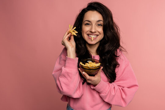 Waist-up portrait of a joyful lady chewing French fries in front of the camera on the pink background. Fast food consumption concept