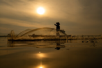 Silhouette of fisherman at sunrise, Standing aboard a rowing boat and casting a net to catch fish...