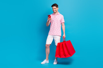 Full body portrait of positive guy use smart phone hold online shop bags isolated on blue color...