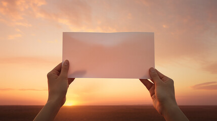 A hand holds a blank white sheet of paper standing against a sunset background. Blank white card for design mockup