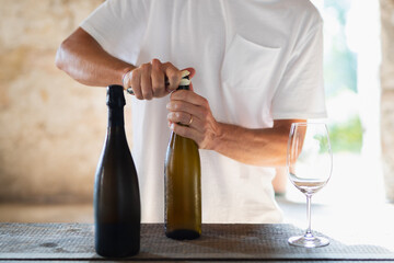 Un ragazzo con maglietta bianca stappa il vino. DUe bottiglie di vino senza etichetta perfette per mockup in ambiente luminoso.