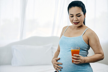 Asia Pregnant Woman Savoring Fresh Orange Juice glass sitting in bedroom with copy space; Nurturing a Healthy Pregnancy