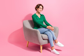 Full length body size photo of smiling girl sitting in grey chair wearing casual green shirt denim jeans isolated on pink color background