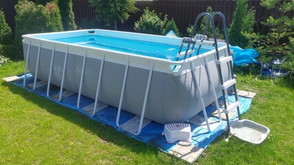 The gray rectangular frame swimming pool stands on a grassy lawn. The water is cleaned with a filter. Next to the metal fence there are thuja trees. The pool is made of PVC. Sunny summer weather