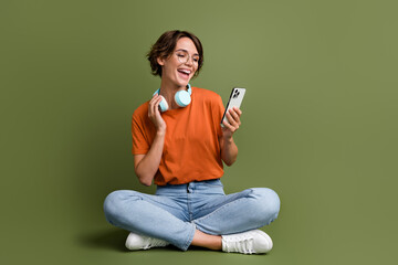 Full length photo of positive girl dressed t-shirt in eyewear sit connecting headphones to smartphone isolated on green color background