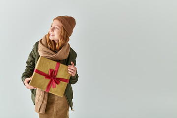 Fototapeta premium smiling girl in trendy winter outfit with knitted hat holding Christmas present on grey backdrop