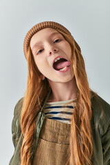 preteen girl in knitted hat and outerwear sticking out tongue and looking at camera on grey backdrop