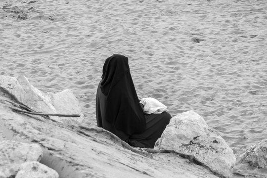 Algerian woman from behind sitting on huge rocks in the Sablettes beach and wearing a black djellabah. Black and white picture.