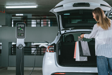 Young woman travel with EV electric car to shopping center parking lot charging in downtown city...