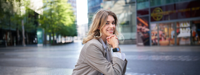 Portrait of woman waiting for an interview, sitting in city center in beige suit, has digital...