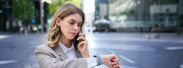 Serious businesswoman checking time on watch and calling person on mobile phone, waiting for...