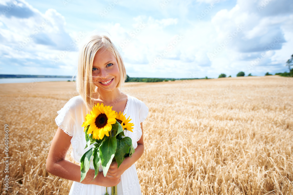 Wall mural Happy woman, portrait and flowers in nature countryside on grass or wheat field with cloudy blue sky. Female person or blonde smile with sun flower, plant or blossom in sunshine, travel or vacation