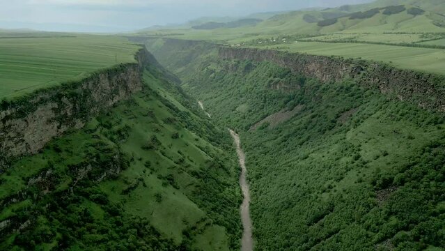 Deep rocky canyon with steep walls. At the bottom is the Dzoraget River near Gnevank Monastery in Armenia. Above is the village of Kurtan against an overcast sky. Drone video. Fly forward.