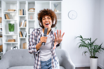 Happy African American woman in wireless headphones using remote control as mic, singing favorite songs at home during coronavirus quarantine
